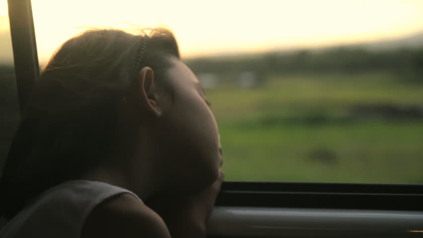 Young girl looking out from car window. Sunset at the background.