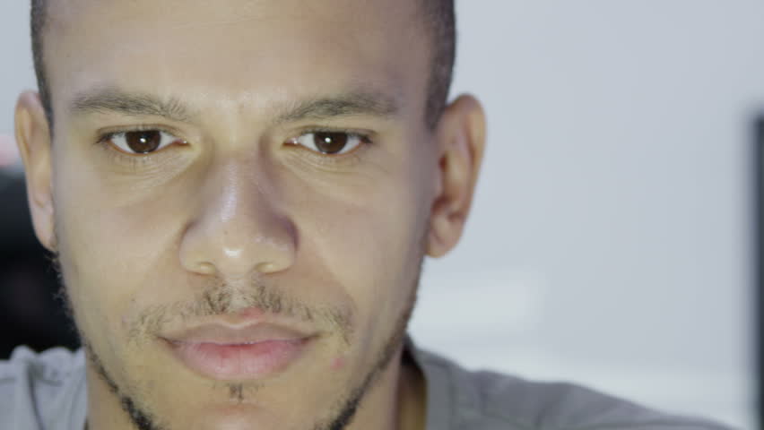 Close up of a very tired looking young man who is working on a computer late at night. He gives a small smile at something he sees on the screen.