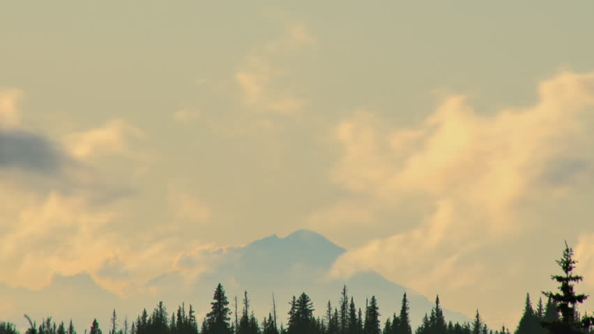 Fast flowing cumulus clouds zooming past Mt. Redoubt in the late afternoon, a silhouetted treeline in the foreground. 