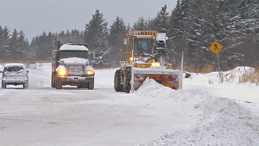Snow blower on an excavator paired with a dump truck to clean plow piles on snowy city street.