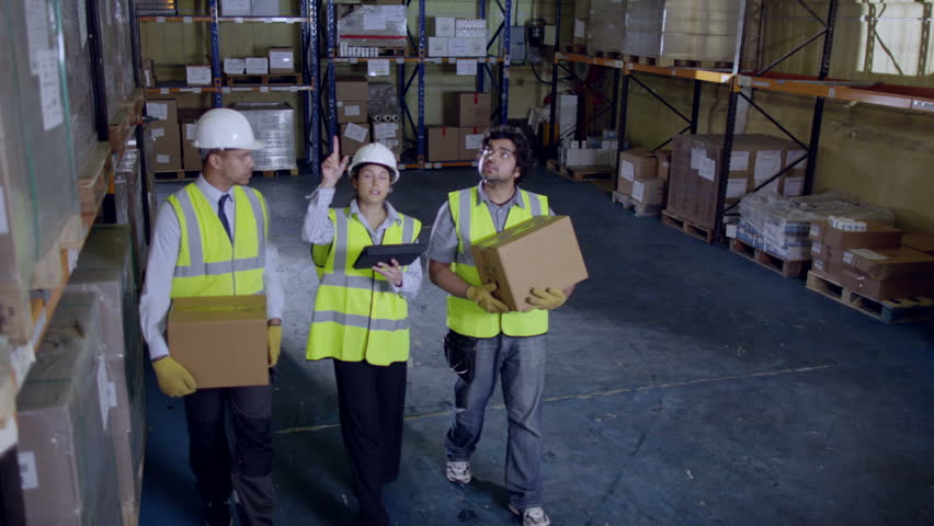 3 workers are walking along the aisle of a warehouse and checking their inventory. Two workers are carrying boxes and their female manager is giving them instructions. Slow motion.