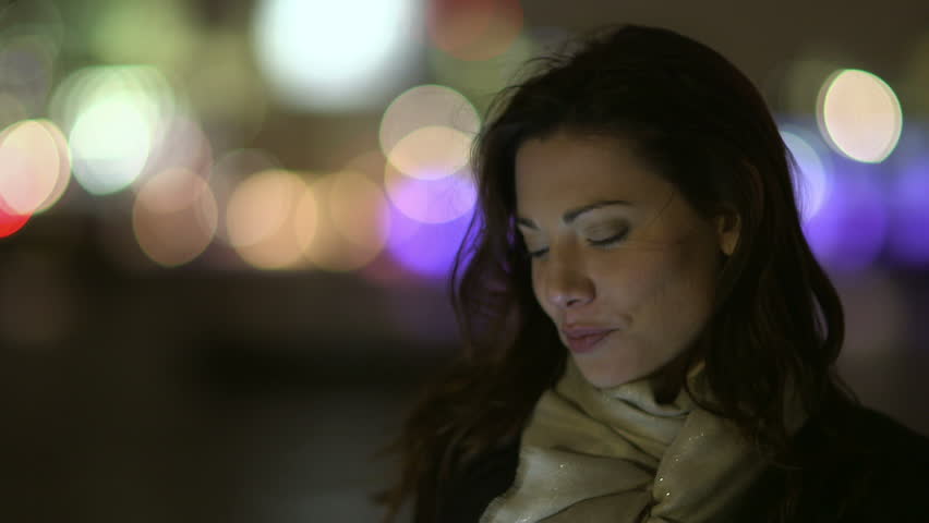 A beautiful young professional woman is having a phone conversation, outdoors in the city at night. She is smiling and the bright lights of the big city can be seen in the background behind her.