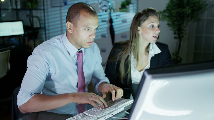 A businessman and businesswoman are working late at night in a room full of computers. They are looking at the screens in front of them and discussing their work