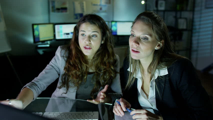 2 attractive female office workers are working late at night, a bank of computer screens can be seen in the background. Slow motion.
