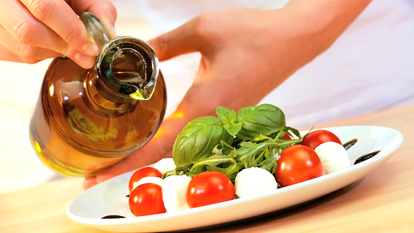 Olive oil being used as a dressing on healthy meal of fresh salad greens, tomatoes & mozzarella cheese balls