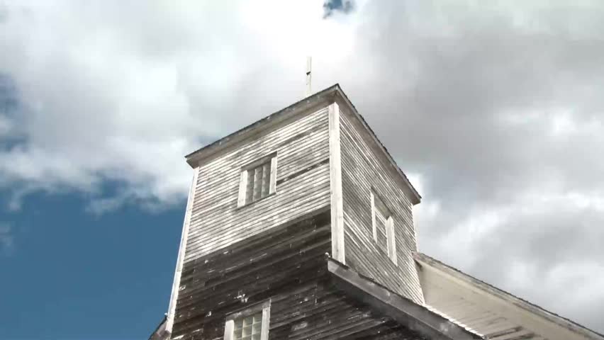Time-lapse of old abandoned church against a blue sky with puffy white clouds rapidly moving past broken steeple.