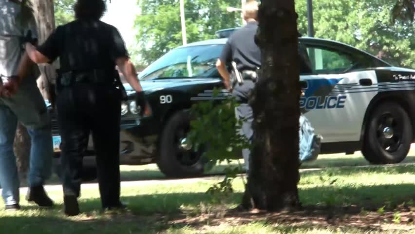 Man appearing to be homeless by a river is arrested by police and then promptly taken to a cruiser.