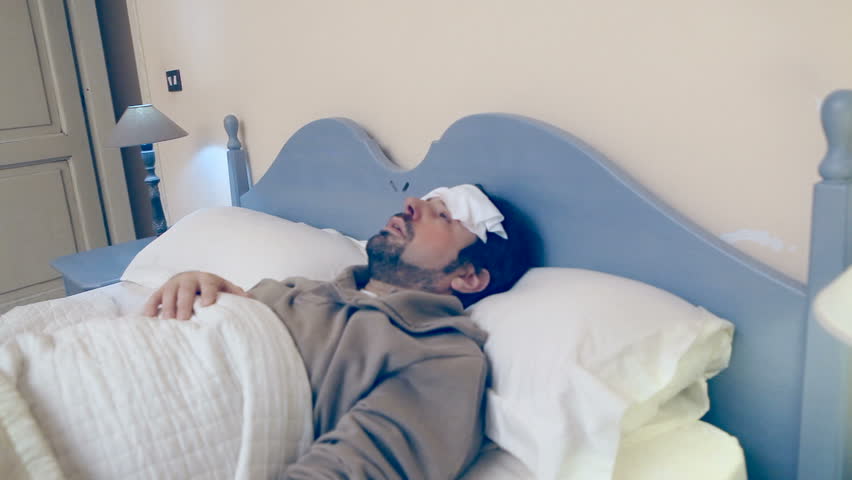 A man lies in the bed, he's got a bad flu / cold / illness. He looks at the thermometer, then he despairs because his body temperature is too high.