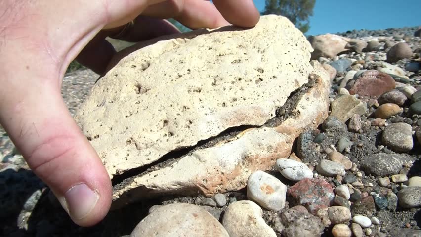 Pale yellow rock is broken in half in sunlight on the banks of a railroad track; hand picks up one half to open middle, and then places it back down.