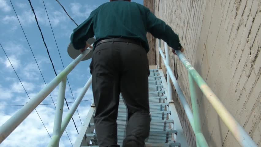 Businessman walks up shaky metal stairs in downtown urban setting with wires and blue sky in background.