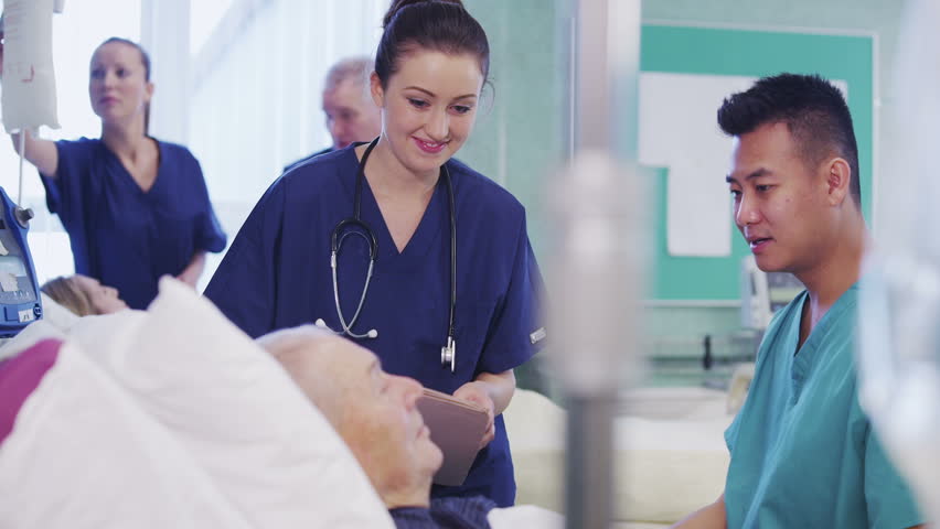 One male and one female nurse attend to an elderly male patient, checking his blood pressure and chatting with him. In the background a nurse and consultant specialist are discussing another patient.