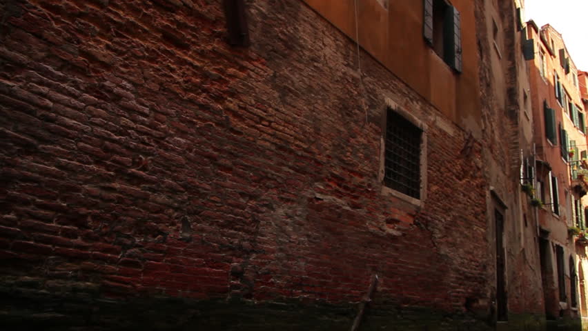 Tracking shot from a gondola gliding along a canal in Venice. The camera is pointed up at the top of buildings, taking in the top floors and roofs as they pass by. 