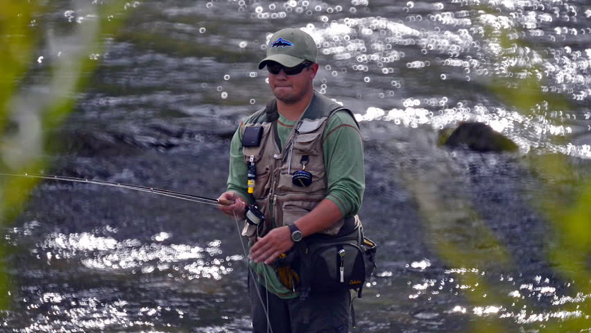 A man in waterproof overalls stands in a river and casts his lure with his rod while he spends his day fly fishing.