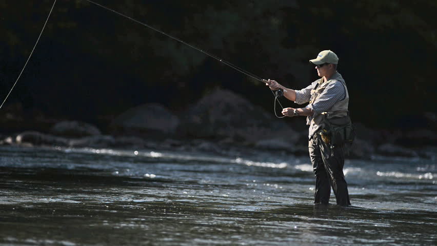 A man in waterproof overalls stands in a river and casts his lure with his rod while he spends his day fly fishing.