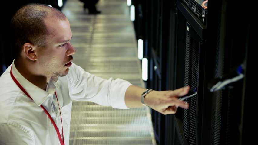 Three people of mixed ethnicity are working in a data center with rows of server racks and super computers. They are walking up and down and checking all of the equipment. 