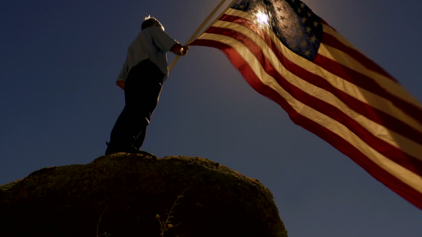 Man, silhouetted, lit by sun, stands on boulder, holds American flag that waves in strong wind, slow motion. 1080p   