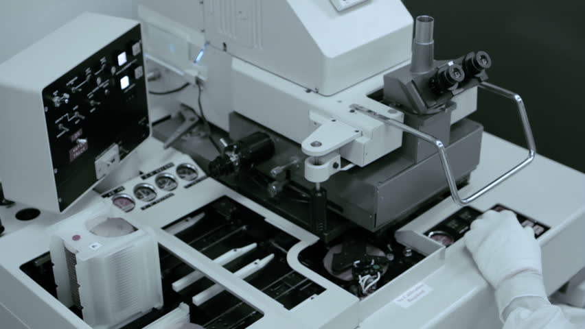 Technician checks part of the process during silicon chip manufacture in a clean room. He is wearing full body white "bunny suit" coveralls to avoid contamination. Over the shoulder view of equipment.