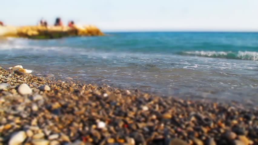 Focus on coastline, pebble and waves of blue sea in motion. Sandy beach, people in background. Tranquil seascape. Beautiful view with scenic landscape. Idea of summer vacations. Relax on beach.
