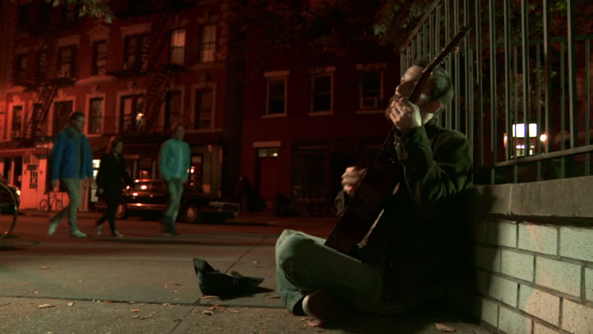 Young man playing a worn-out guitar on the night time streets of Manhattan, NY. Young woman approaches, listens for a while then leaves some money. 