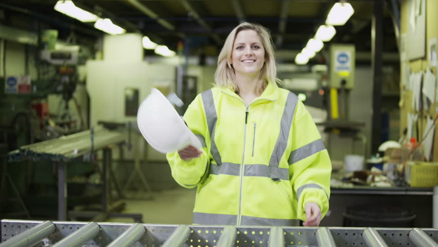 Portrait of a cheerful and friendly female warehouse manager who is wearing a hard hat and reflective clothing. She takes off her hard hat and smiles at the camera.  In slow motion.