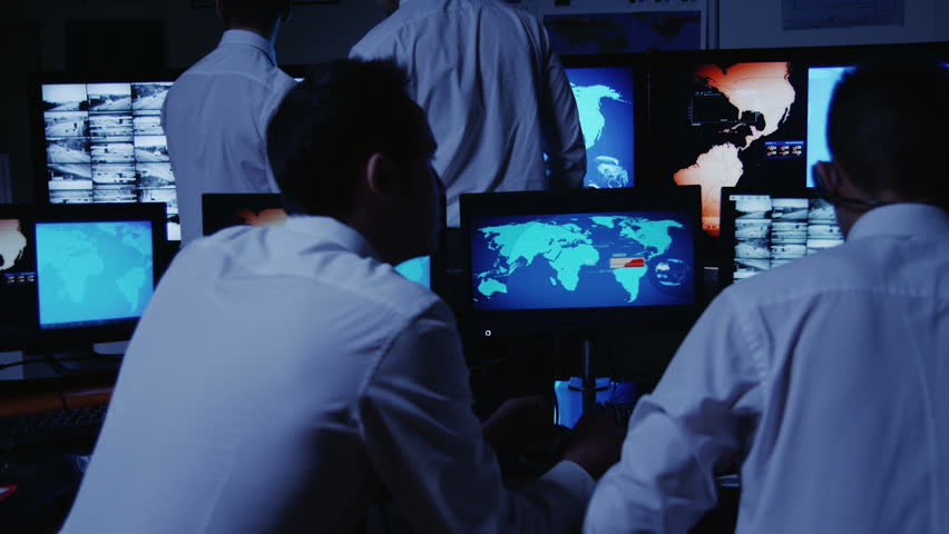 A team of security personnel man the stations within a busy system control room. Could be a weather or power station or air traffic control. It could be a  police/army control facility. Slow motion.