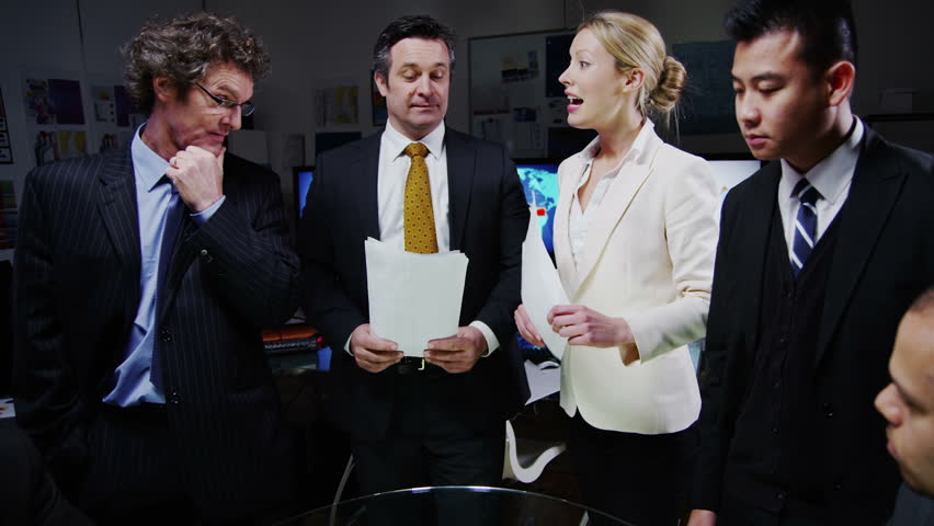 A diverse team of business professionals are having a  late meeting in a dark room full of computer screens. They are looking over sales figures and discussing possibilities for business development.