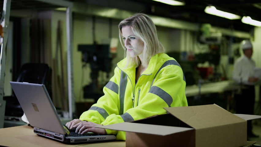 A beautiful female warehouse employee wearing high visibility clothing and a hard hat is working on a laptop computer and checking her stock.