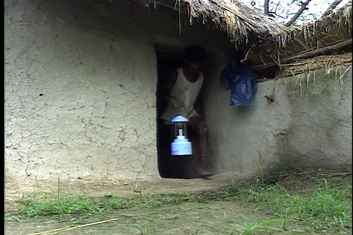 SULTANPUR, INDIA - JULY 28, 1999: Man walks out of house carrying solar powered lamp. Sits down next to solar panel and plugs lamp in to charge.