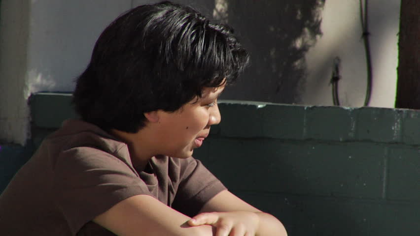 A teenage boy sits on some steps looking around, and eventually puts his head in his arms.