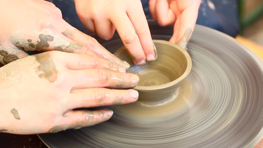 A potters hands guiding a child hands to help him to work with the ceramic wheel 