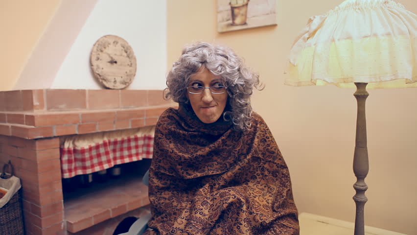 Funny scene of a granny (a dressed up actress) with a scarf, on a rocking chair, secretly drinking in hiding some alcoholic liquid from a flask (a glass bottle). Humorous shot.