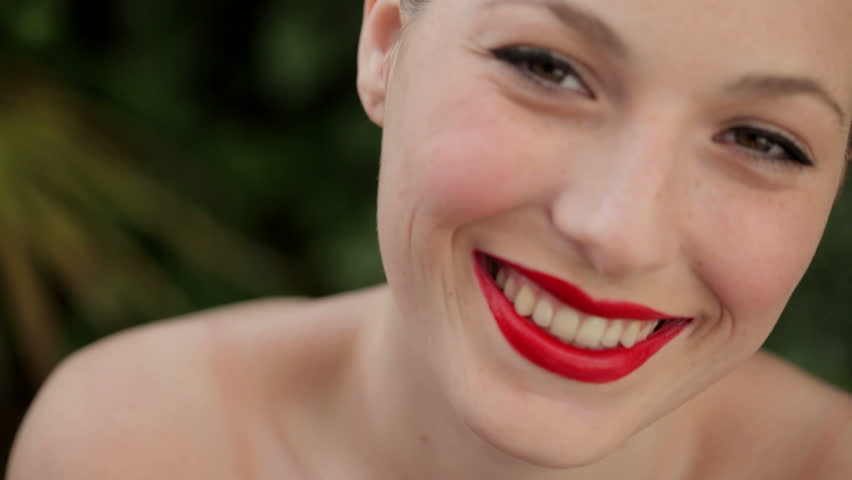 Close up portrait of a young beautiful woman with bare shoulders, wearing bright red glossy lipstick and smiling outdoors, in a green forest background.