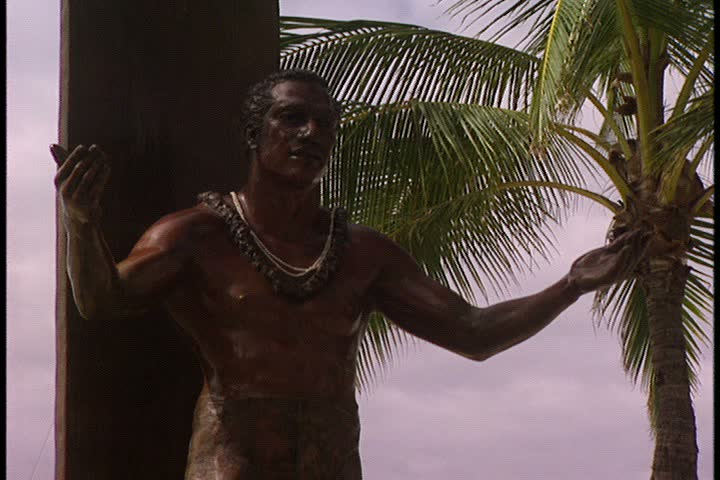 WAIKIKI BEACH, HONOLULU, HAWAII - JANUARY 30, 1997: CU of Duke Kahanamoku (surfer) statue and palm trees in frame then pull out to WS of people on the beach.