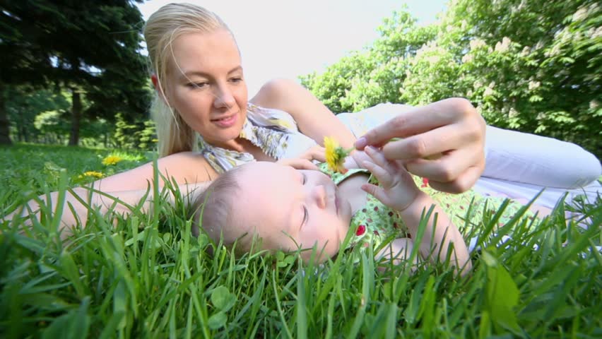 Young mother lies near her baby daughter and they play with yellow dandelion, on grass at sunny summer day in park