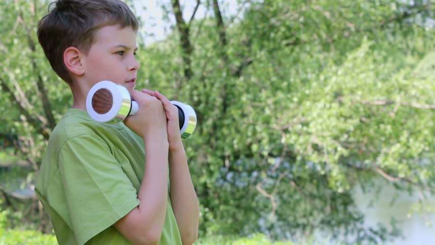 Boy in green t-shirt does exercises with dumbbell in park at sunny summer day