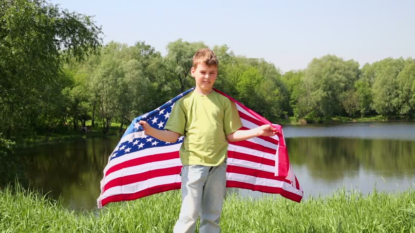 Boy in green t-shirt holds unfolded US flag near pond at sunny summer day