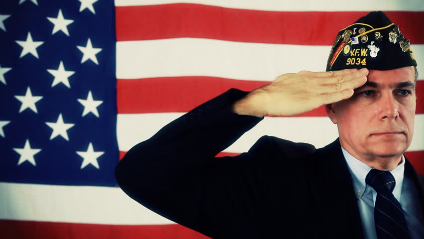 A man in a VFW cap, standing in front of an American Flag, stands ready and then salutes. Dolly Shot.