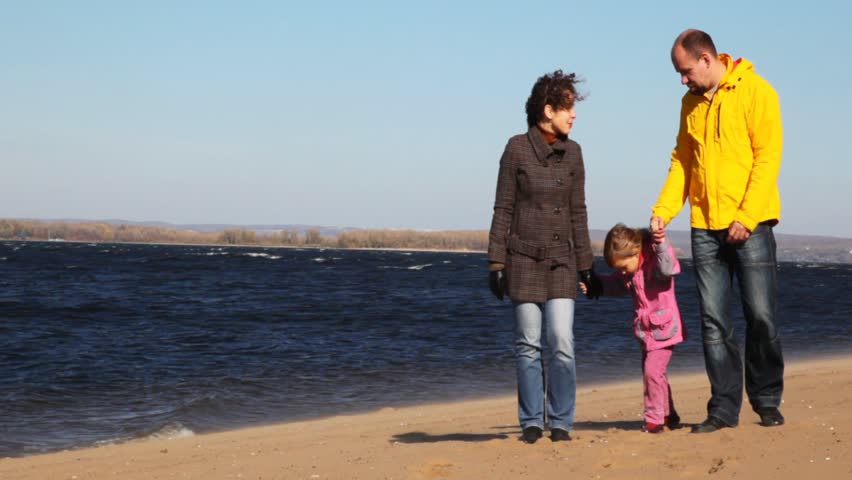 family of mum, daddies and daughter stands on sandy to river bank and keep for hands 