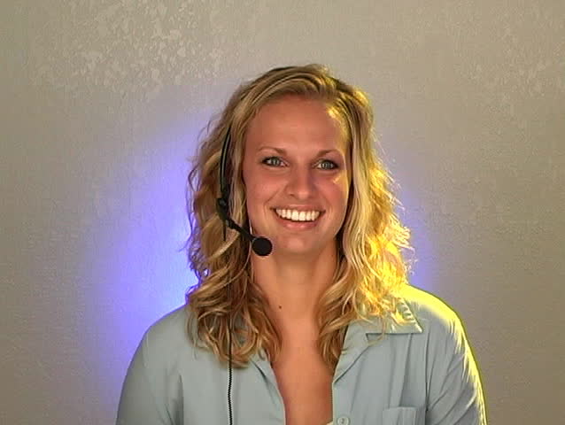 A beautiful young blonde customer service operator wearing a headset telephone microphone wraps up a call and looks at the camera with a bright, warm smile. 