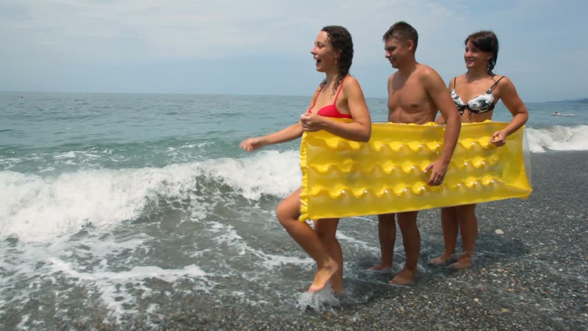 two women and man holds inflatable mattress walking on beach 