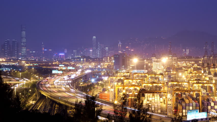 Busy City. Timescape at Night. Hong Kong. Zoom in shot. Corporate buildings at the back. Busy cargo container loading activity in container terminal. Busy traffic across the main highway at rush hour.
