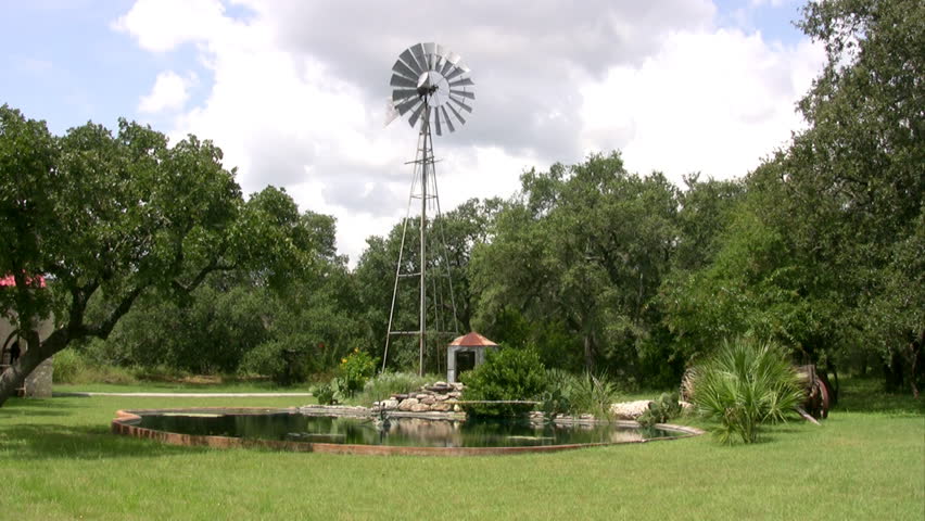 Video closer of a windmill on a south Texas ranch. Spinning in the wind with fluffy clouds in sky. Green ranch field with small stock pond and trees around windmill used to water livestock cattle. 