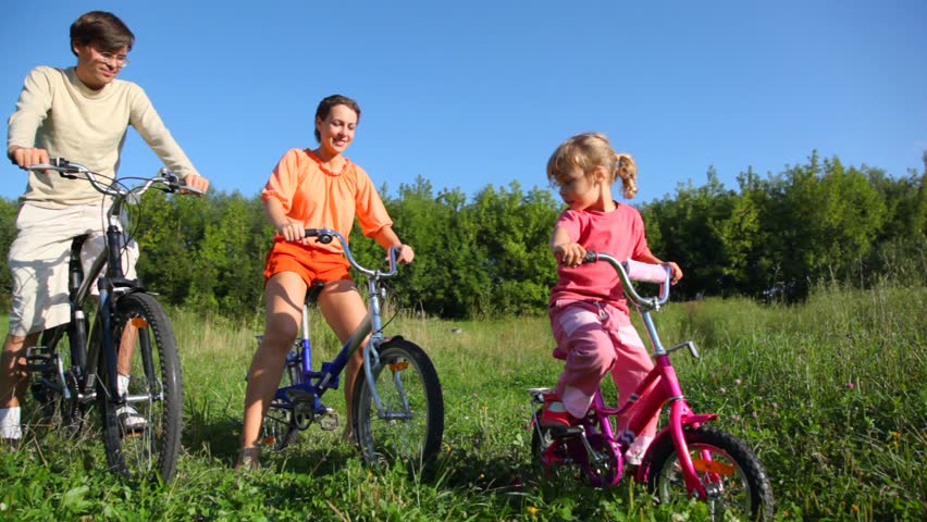 family of three with bicycles in field, mother pushes slightly her daughter 