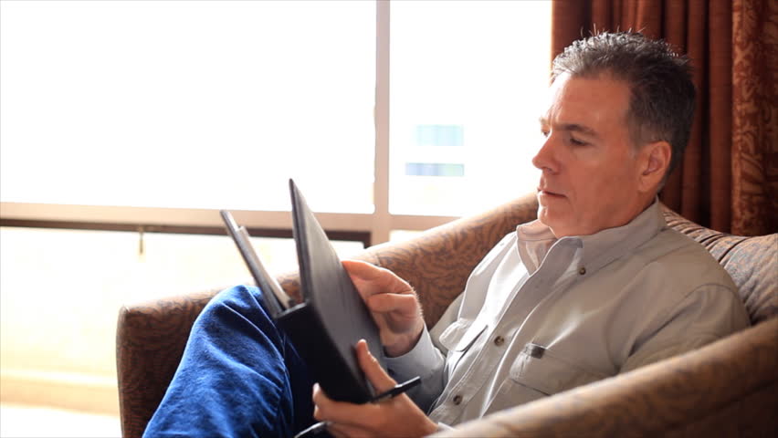 A relaxed man sitting by a big bright window in a large lounge chair writing something in a notebook.