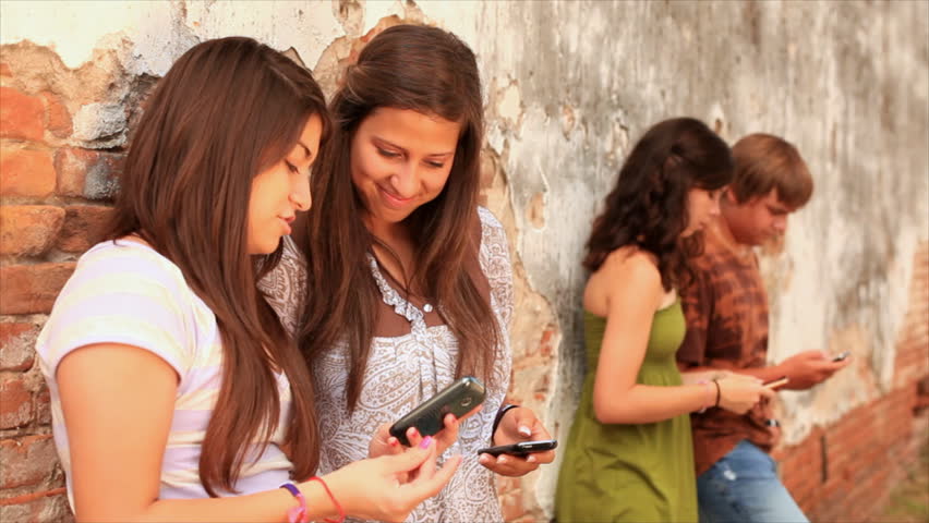 A group of teenagers hanging out by an old brick wall, sharing text messages with each other.