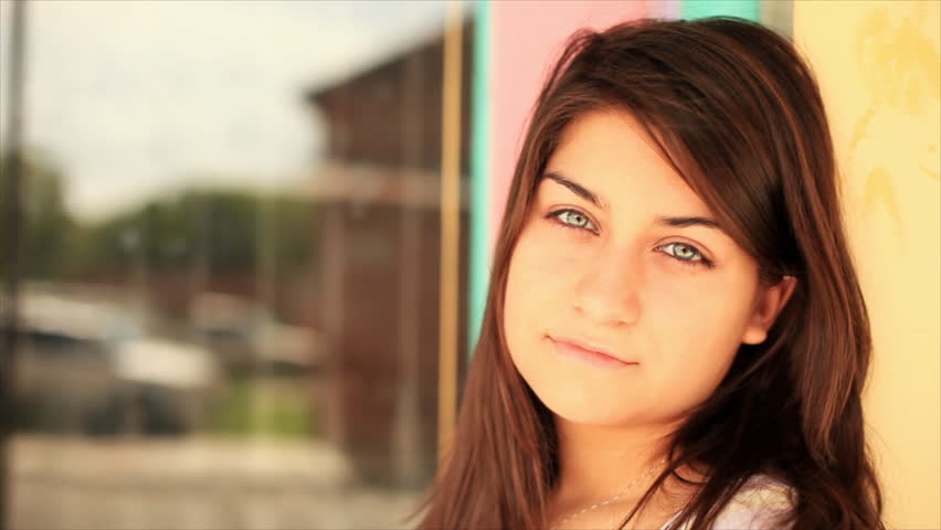 A beautiful brunette girl with piercing eyes gazes at the camera as she leans against a colorfully painted wall.