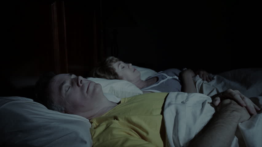 A mature woman sleeping next to her husband is awakened by heartburn, which she then takes medication for.