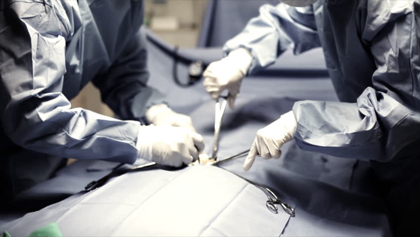 Two surgeons in full surgery garb, using surgical instruments, operate on a patient in an operating room. Dolly or tracking shot.