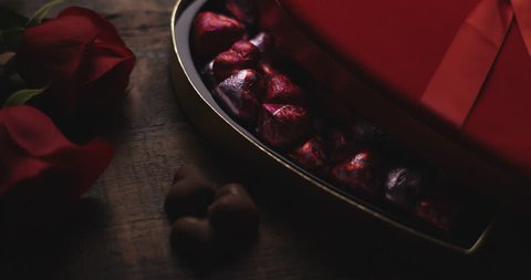 Heart shaped box of chocolates in wrappers on wooden table with roses. Light slowly fades up.