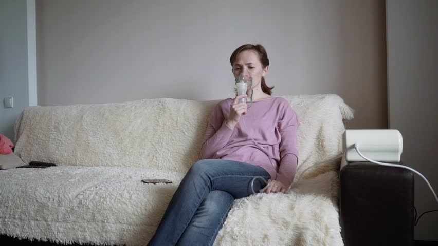 A Woman Sits On The Couch At Home And Inhales Through An Inhaler Inhalation Mask A Woman Sits On The Couch At Home And Inhales Through An Inhaler Inhalation Mask
